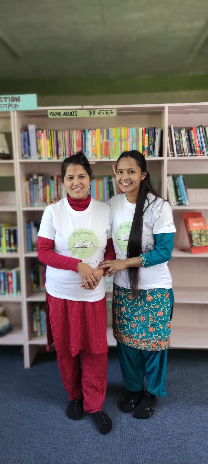 Zwei weibliche Bibliothekarinnen vor einem Bücherregal, beide tragen ein T-Shirt mit dem Sapana Logo